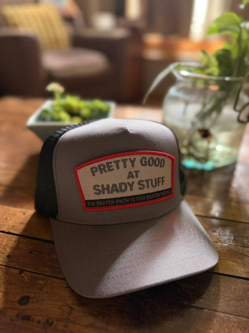 Pretty Good at Shady Stuff trucker hat by Rogue Dynamics, gray and black with red-bordered patch, on a wood table